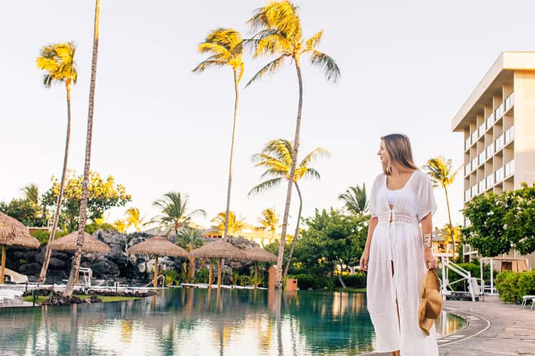 Woman in white dress at tropical resort pool with palm trees, thatched umbrellas, and hotel building background