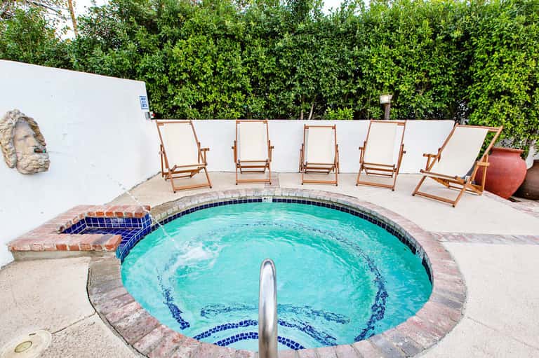 Circular turquoise pool surrounded by wooden lounge chairs and white walls with tropical greenery backdrop