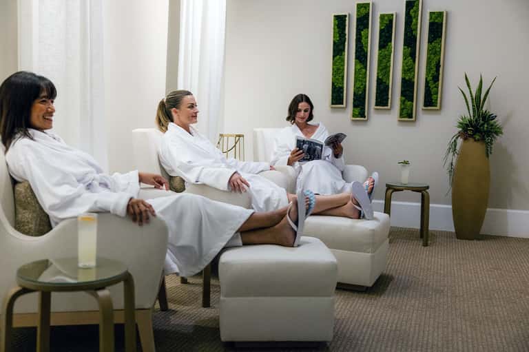 Spa relaxation room with three women in white robes on cream furniture with plant decor