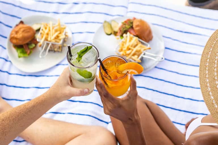 Hands holding refreshing beverages with burgers and fries on striped beach towel