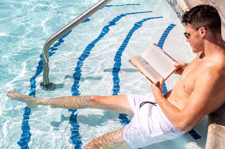 Man reading by swimming pool edge with blue lap lanes, relaxing in white swim trunks at resort pool