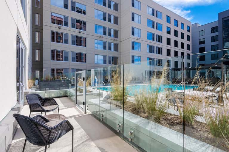 Modern hotel pool area with glass safety railings, lounge chairs, decorative grasses, and multi-story buildings