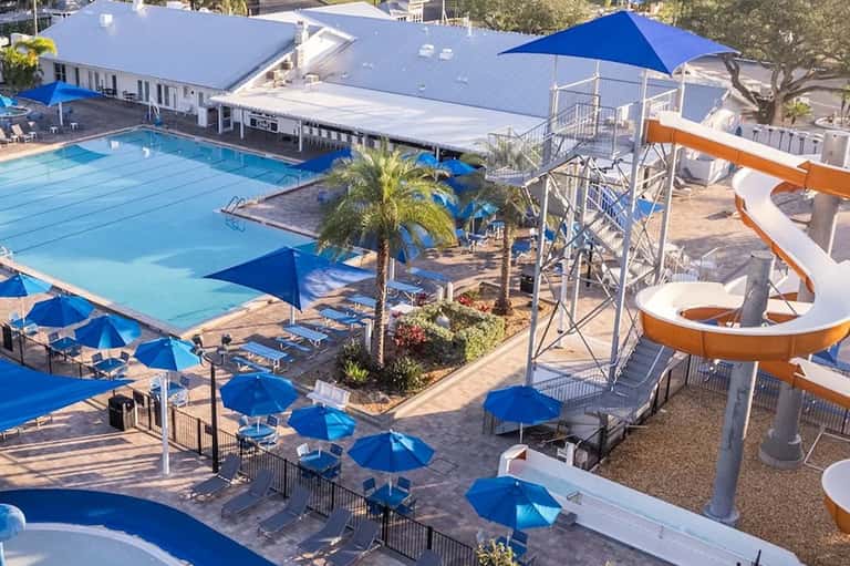 Aerial view of resort Olympic pool with blue umbrellas, water slide, and palm trees