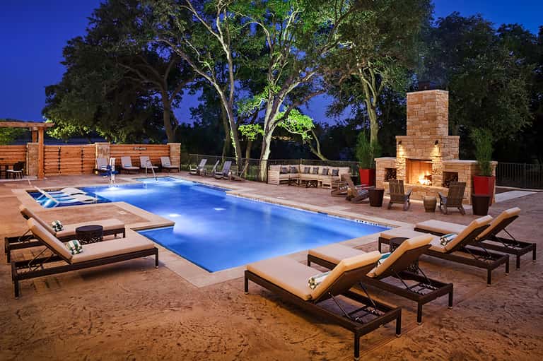 Illuminated resort pool at night with lounge chairs, fireplace, and mature trees