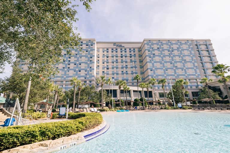 Large resort hotel with curved pool, palm landscaping, lounge chairs, and modern architecture by waterfront