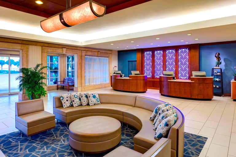Modern hotel lobby with curved seating, ocean view windows, wood paneling, and purple accent lighting