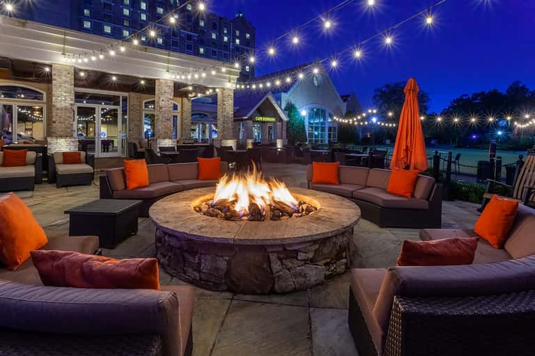 Outdoor fire pit seating area at night with brown sofas, orange pillows, and string lights