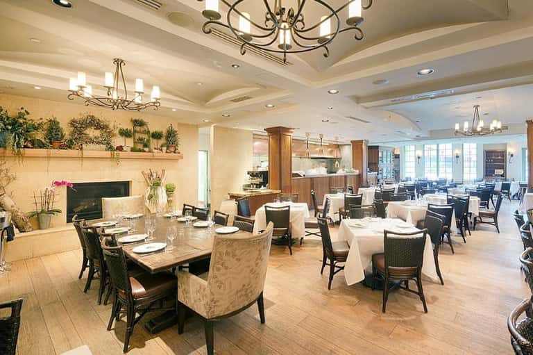 Fine dining restaurant with white tablecloths, fireplace, decorative plants, and high ceiling with chandeliers
