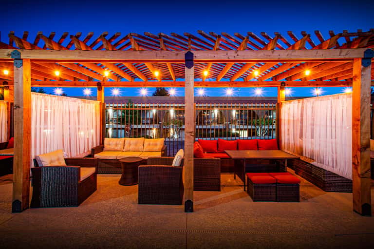 Covered poolside lounge area at dusk with wooden pergola, ambient lighting, and upholstered seating