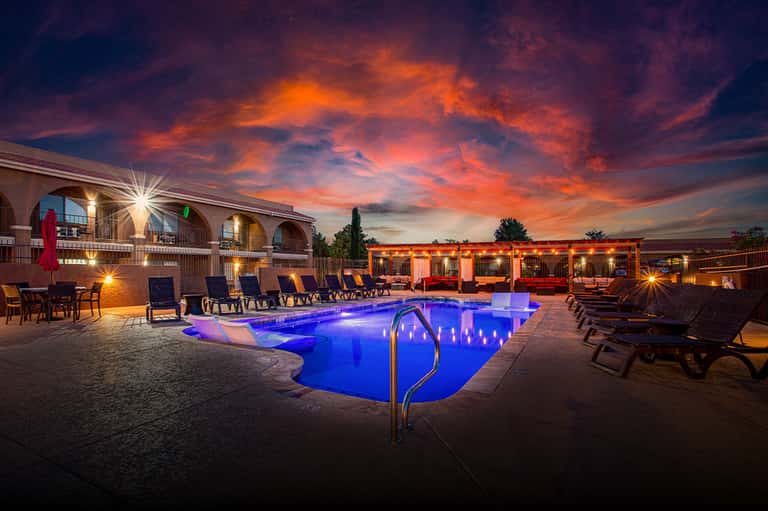 Resort pool glowing blue at dusk with lounge chairs, umbrellas, and dramatic orange sunset sky