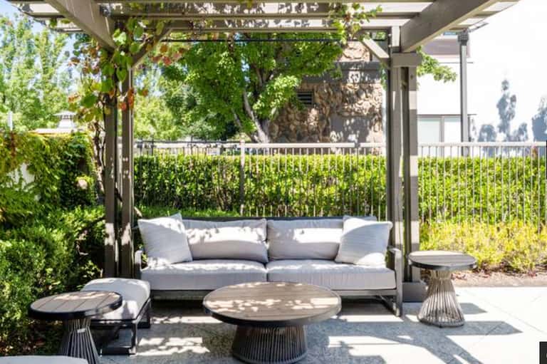 Modern pergola with gray sofa, round coffee table, and lush green privacy screening