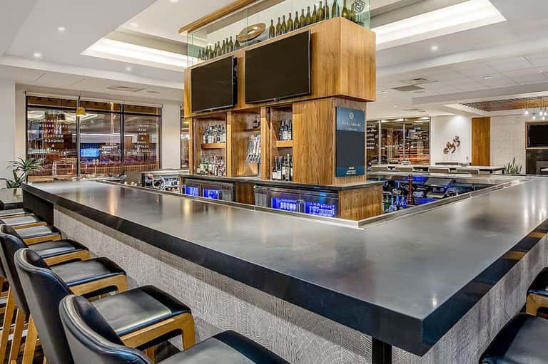 Modern hotel bar with wooden display cabinet, metal countertop, bar seating, and multiple beverage selections