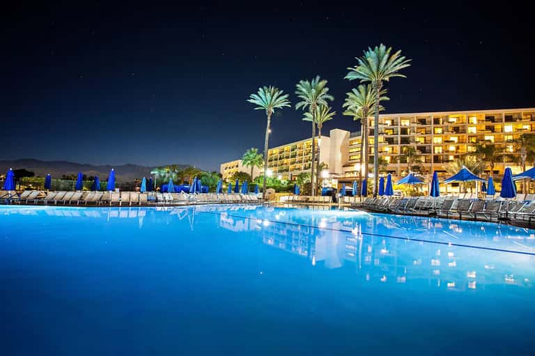 Illuminated resort pool at night with blue umbrellas, palm trees, and lit hotel building