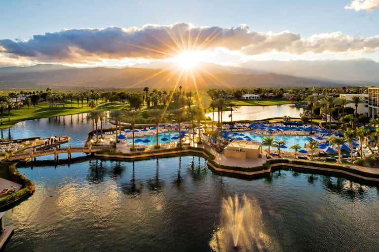 Resort pool complex at sunset with palm trees, blue loungers, fountain, and mountain backdrop