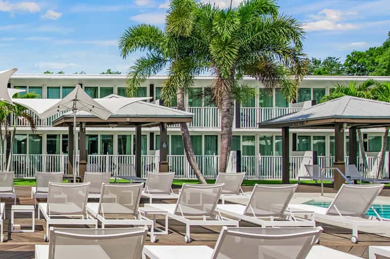 Seaside resort pool deck with white lounge chairs, palm trees, white umbrellas, and building with green shutters