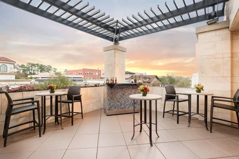 Rooftop bar terrace at dusk with high-top tables, bar counter, pergola, and city skyline views