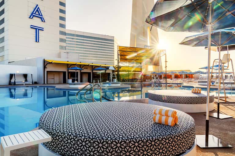Resort pool deck with modern loungers, umbrellas, water features, and hotel building