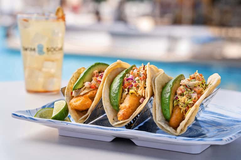 Three fish tacos with avocado and coleslaw on decorative plate by pool area