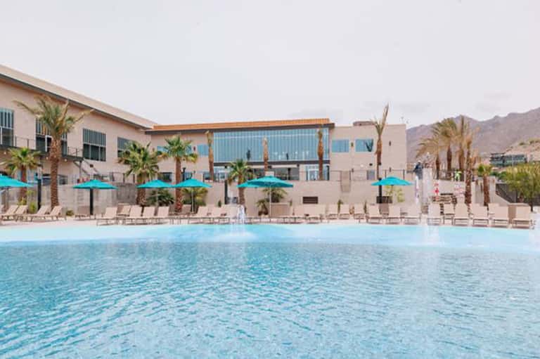 Resort pool with turquoise water, palm trees, blue umbrellas, and modern beige buildings