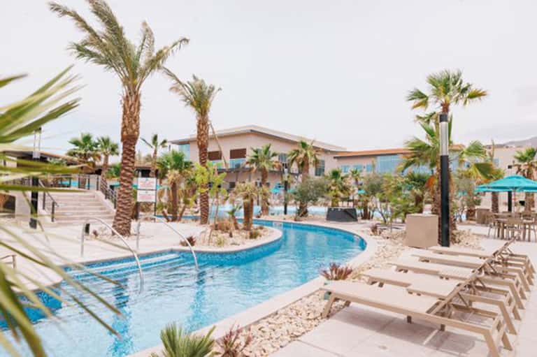 Resort pool area with curved lazy river, palm trees, lounge chairs, and main building in background