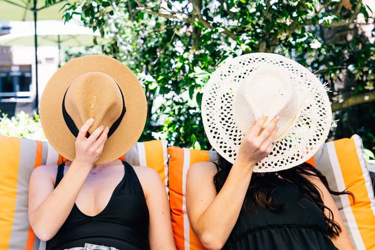 Two women in pool loungers wearing straw hats and holding them in front of their faces