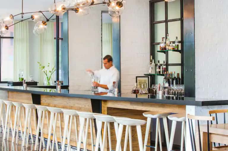 Contemporary bar with long counter, white stools, pendant lighting, and bartender in background