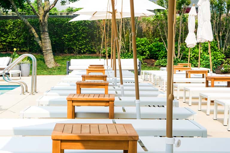 Poolside lounge area with white cushioned benches, wooden end tables, white umbrellas, and palm trees