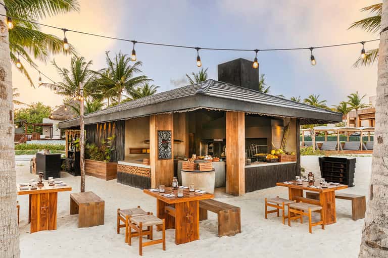 Rustic beachside restaurant with wooden furniture, palm trees, and string lighting at dusk