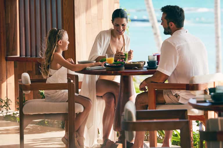 Family dining at outdoor table overlooking ocean with palm trees and tropical breeze