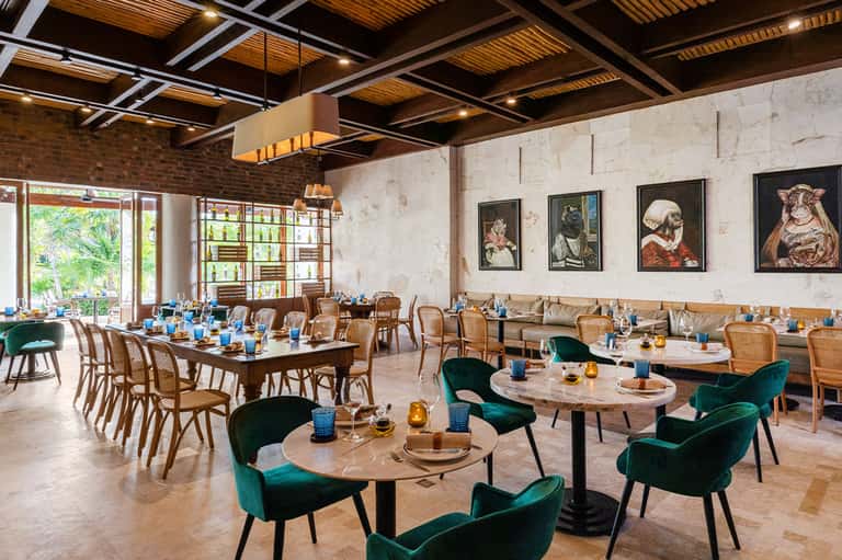 Modern dining room with green velvet chairs, wooden tables, exposed ceiling beams, and natural light