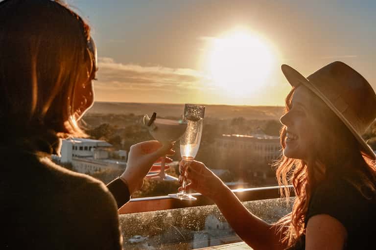 Two women toasting champagne glasses at sunset overlooking coastal city landscape