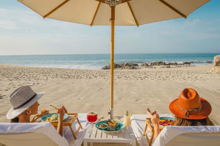Two women dining under beige umbrella on sandy beach with turquoise ocean and rocky coastline backdrop