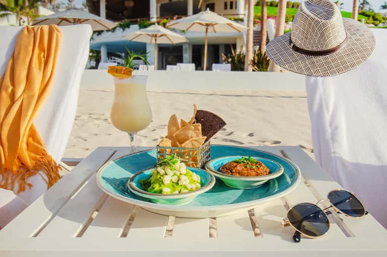 Beach dining setup with fresh salad, cocktail, chips, straw hat, and ocean view backdrop