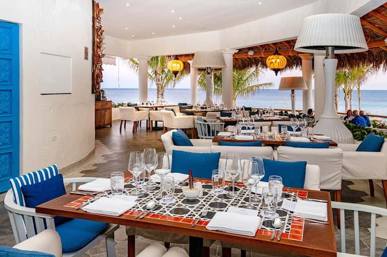 Beachfront restaurant with white columns, blue cushioned seating, palm trees, and ocean views