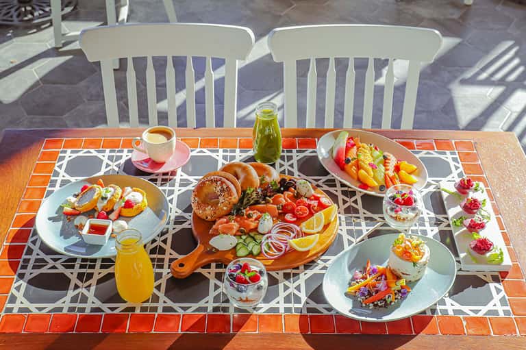 Outdoor dining table with colorful breakfast spread, fresh juices, and pastries on terracotta tile patio