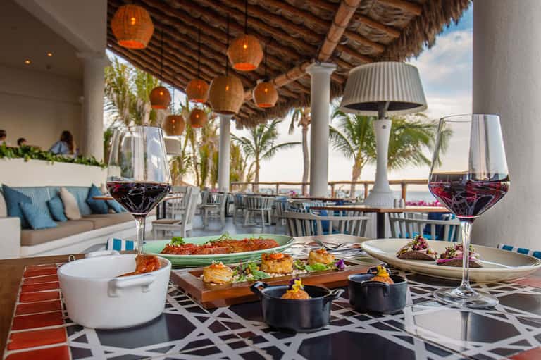 Beachfront dining table with wine glasses, gourmet plated dishes, hanging lanterns, and ocean views from covered terrace