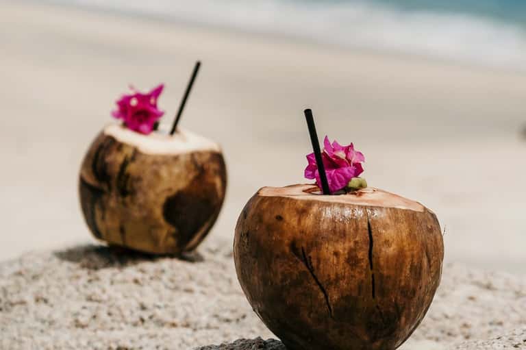 Two coconut drinks with pink flowers on white sandy beach with ocean backdrop