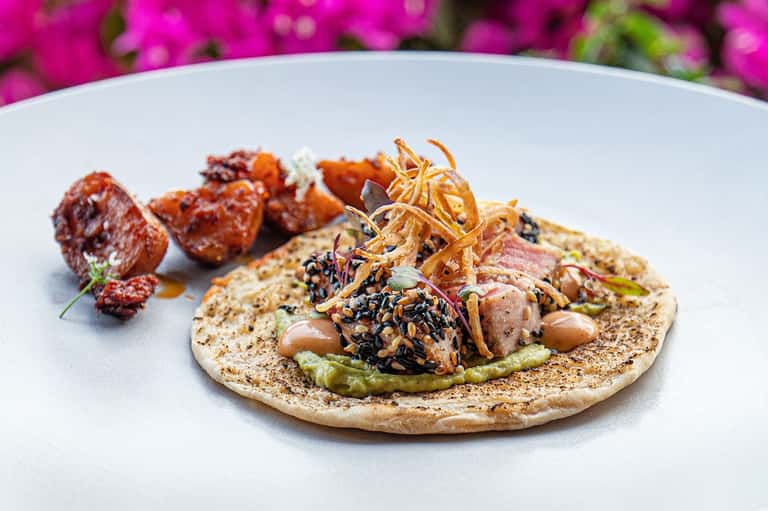 Gourmet plated dish with seared meat, vegetables, black seeds, and magenta bougainvillea flowers backdrop