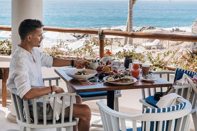 Man dining on beachfront terrace with fresh food, ocean view, and rocky coastal landscape