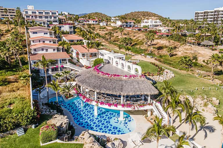 Aerial view of beachfront resort with pool, thatched roof cabana, and hillside villas