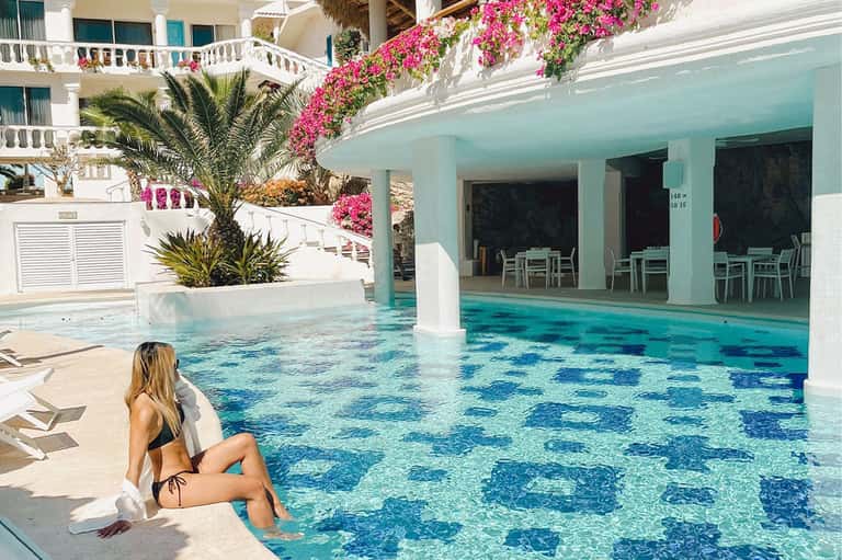 Woman relaxing by crystal-clear pool with pink flowers, palm trees, and white building