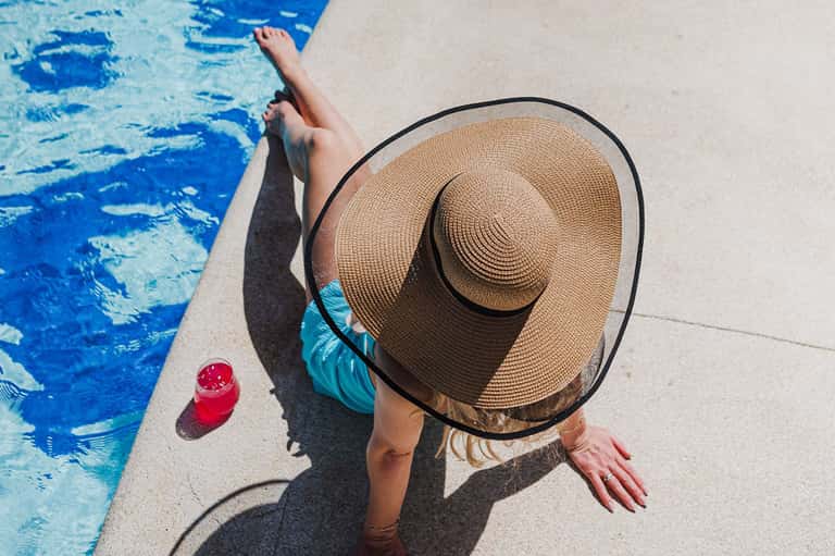 Woman in wide-brimmed hat relaxing poolside with cocktail and turquoise water