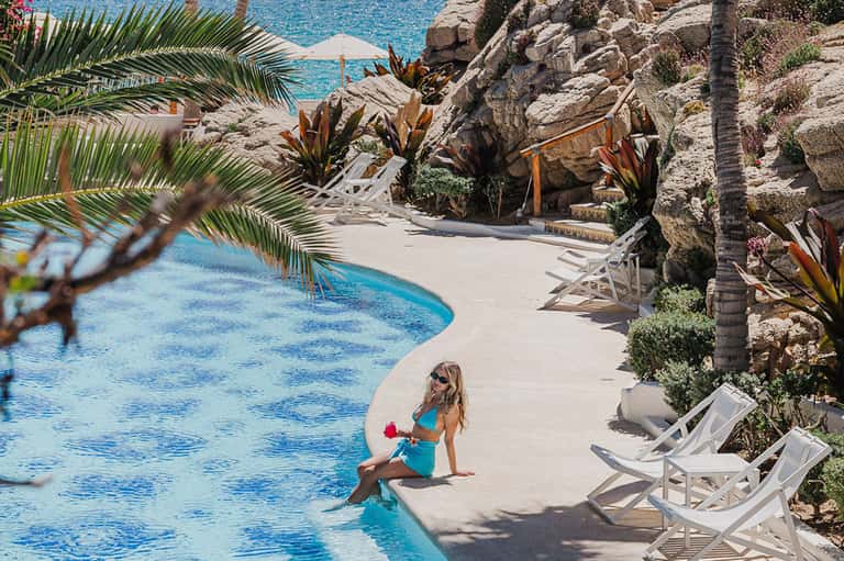 Resort guest in turquoise swimsuit sitting at edge of curved pool surrounded by palm trees
