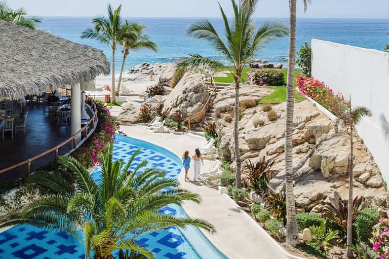 Resort pool area with thatched-roof structures, rocky coastline, palm trees, and ocean view from elevated location