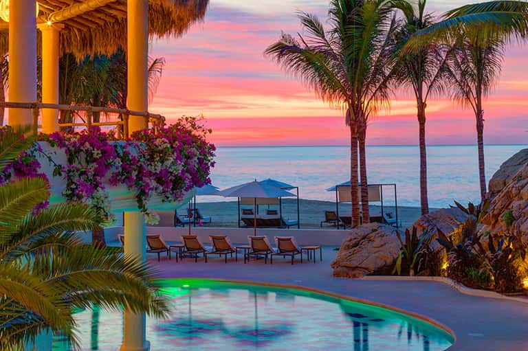 Resort pool at sunset with palm trees, pink sky, and ocean backdrop