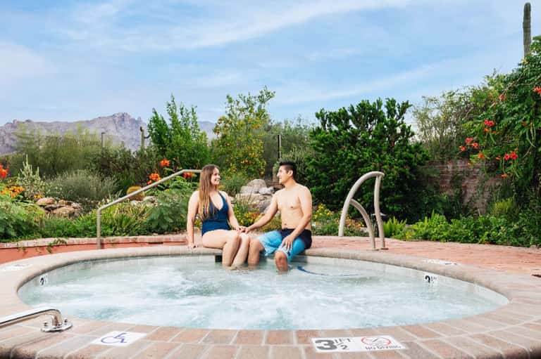 Couple sitting at edge of hot tub with desert mountains, flowering plants and vegetation visible in background