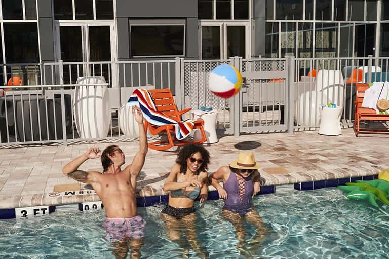 Family playing in shallow resort pool with white fence, colorful beach chairs visible