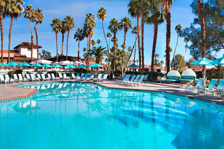 Resort swimming pool with turquoise water, palm trees, lounge chairs, and cabanas under blue sky