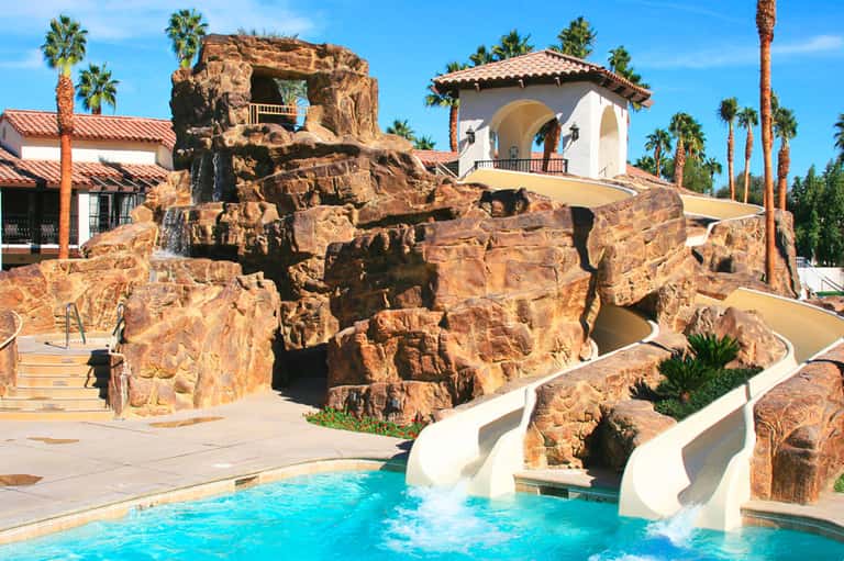 Resort pool with white waterslides, rocky formations, palm trees, and Spanish-style buildings under blue sky
