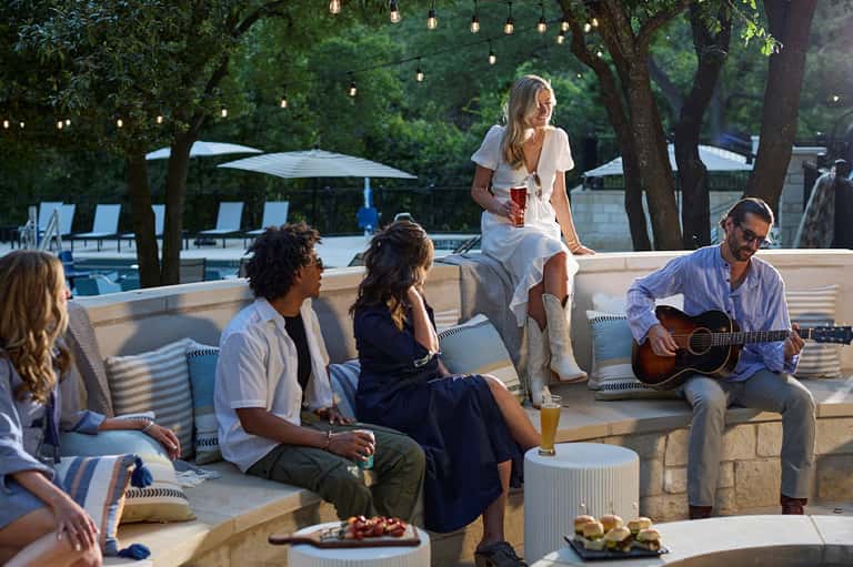 Guests relaxing on outdoor seating by pool with hanging lights, trees, and one playing guitar